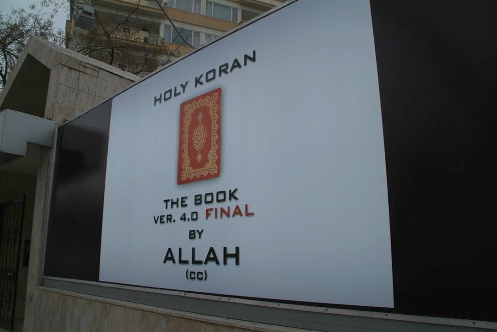 لوحة بيضاء على نظام لوحات ⁦XLight⁩: عبارة "⁦HOLY KORAN⁩" في الأعلى، وصورة كتاب بغلاف أحمر في الوسط، وعبارة "⁦THE BOOK VER. 4.0 FINAL BY ALLAH⁩" في الأسفل — أنقرة، مارس ⁦2006⁩