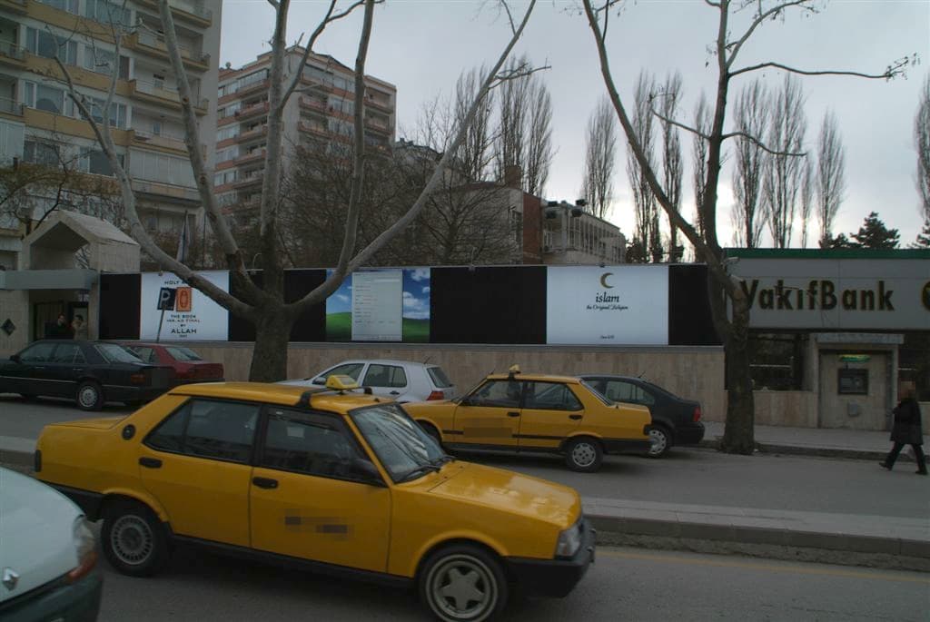 Ankara'da cadde üzerindeki XLight pano duvarının geniş görünümü: üç beyaz zeminli pano, önünde park etmiş taksiler ve araçlar, sağda banka şubesi — Mart 2006